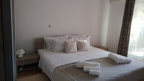 Una cama bien hecha con sábanas blancas y dos almohadones decorativos, una lámpara de noche y una ventana con cortinas.