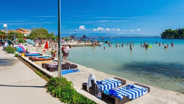 Une plage avec une eau d’un bleu limpide, des gens qui se baignent et qui prennent des bains de soleil, et une jetée en arrière-plan.