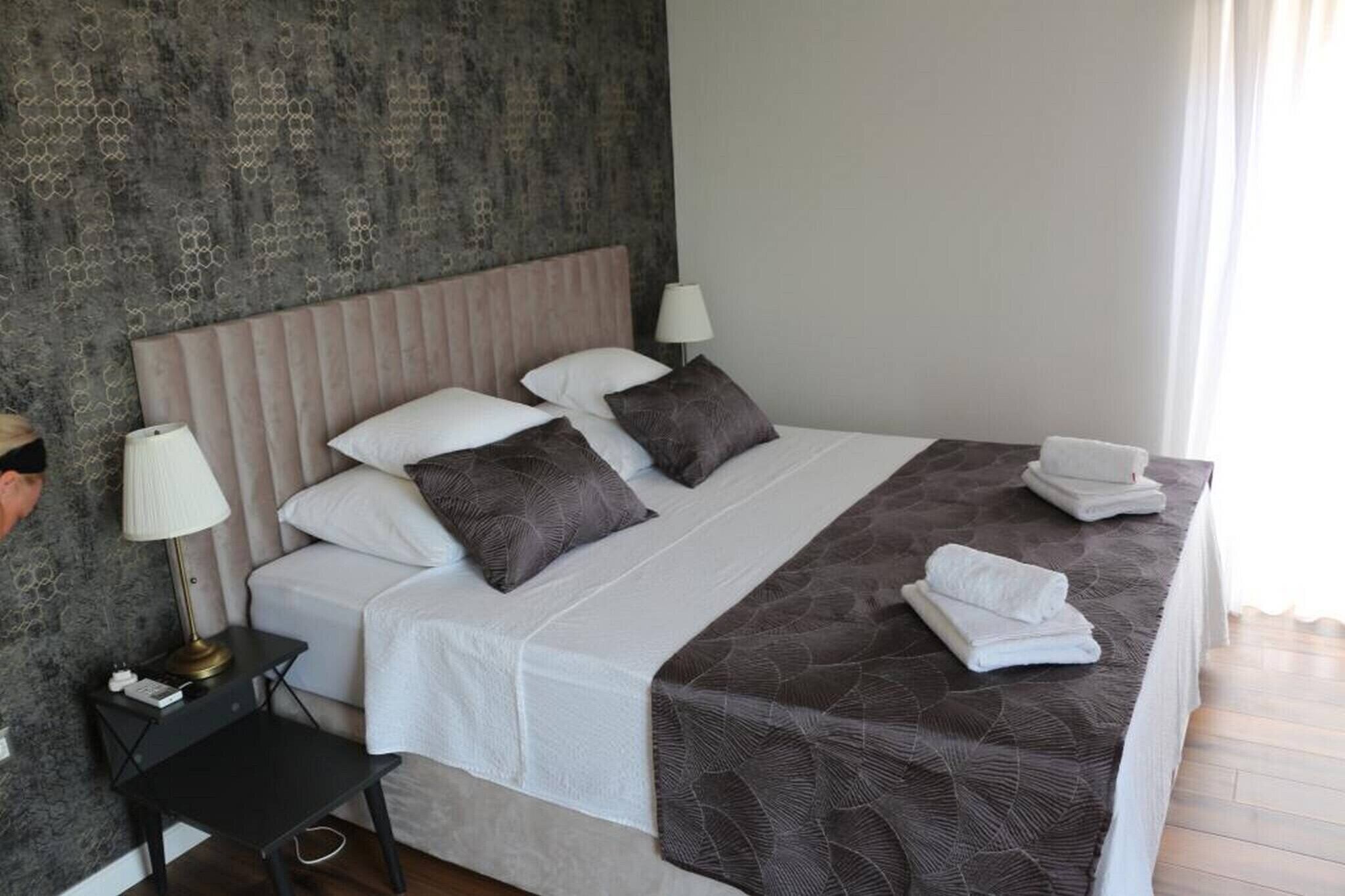A neatly made bed with white linens and a brown patterned bedspread, a bedside table with a lamp, and a textured wall behind the bed.