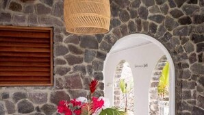 Una habitación con paredes de piedra, una lámpara colgante de mimbre, un carrito de madera con flores y una silla blanca con cojín.