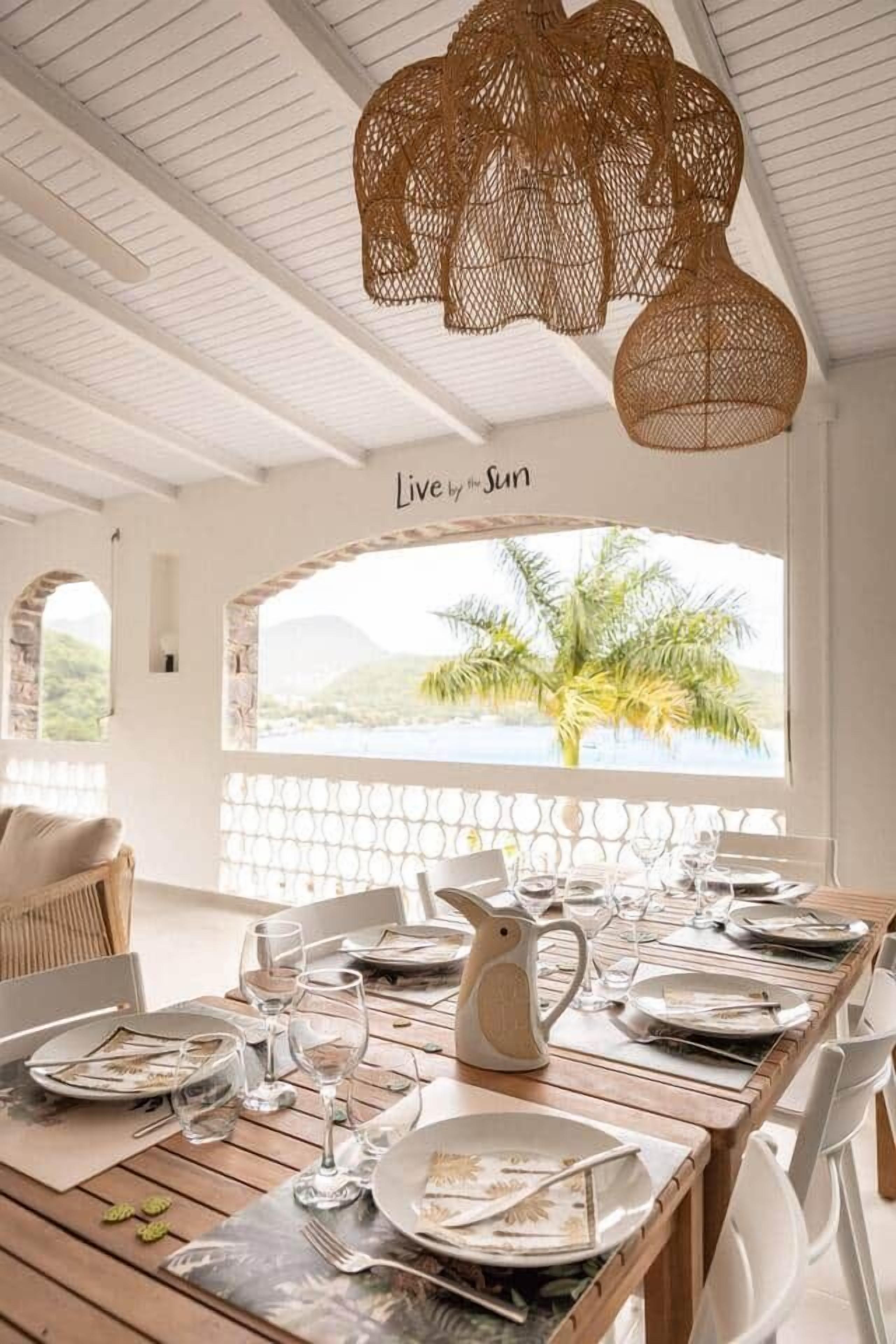 A dining area with a table set for four, a wicker chandelier, and a view of palm trees through a window.