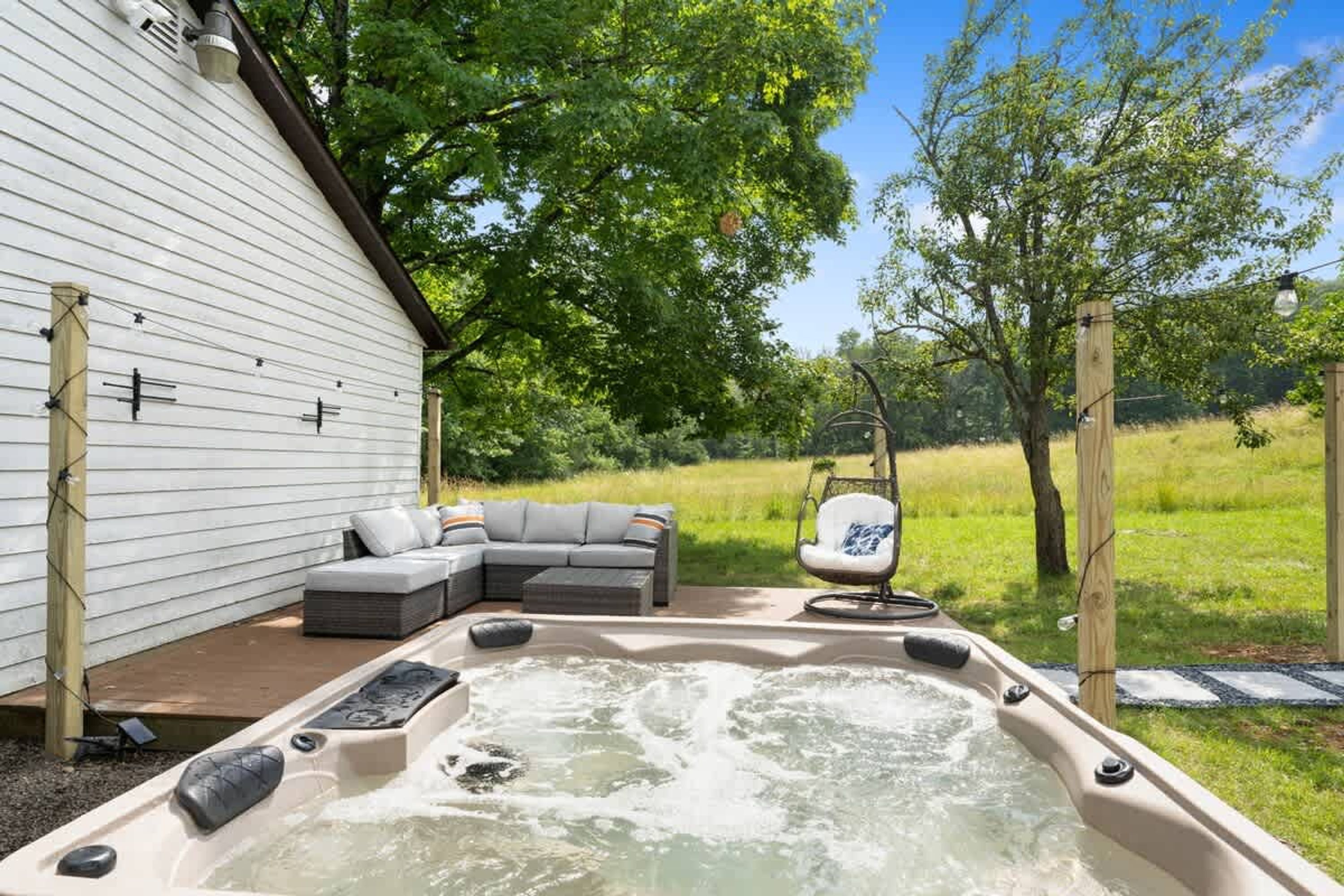 A hot tub with a wooden deck, a gray sofa, and a hanging chair in a garden setting.
