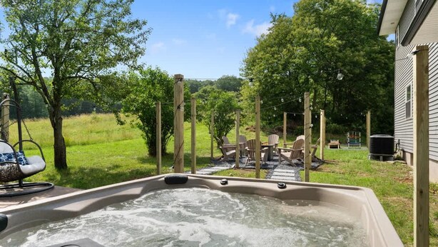 A hot tub with a wooden deck and chairs, surrounded by a grassy area and trees.
