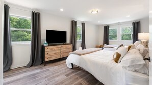 A modern hotel room with a large bed, a flat-screen TV, a wooden dresser, and a window with curtains.