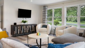 A modern living room with a flat-screen TV mounted on the wall, a round coffee table with decorative items, and a large window offering a view of greenery outside.