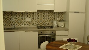 A compact kitchen with white cabinets, a gas stove, an oven, and a sink. There is a dining table with a placemat and a bowl of fruit.