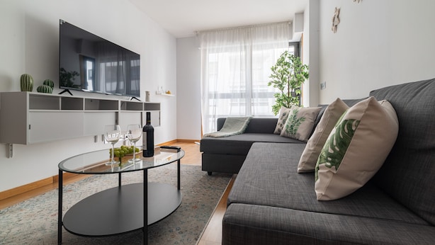 Un salon moderne avec un téléviseur à écran plat fixé au mur, une table basse ronde en verre, un canapé gris agrémenté de coussins décoratifs et une grande fenêtre ornée de rideaux légers.