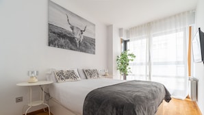 Une chambre à coucher avec un grand lit, une table de chevet, une lampe, une plante et une fenêtre avec des rideaux.