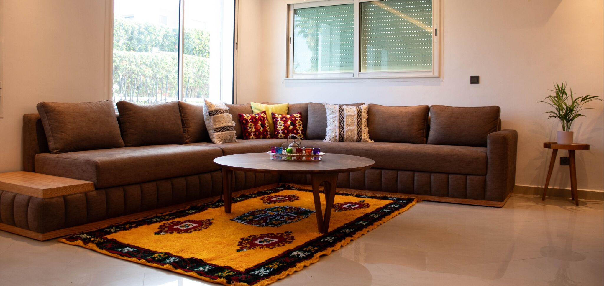 A living room with a brown sofa, a round coffee table with colorful items, and a patterned rug.