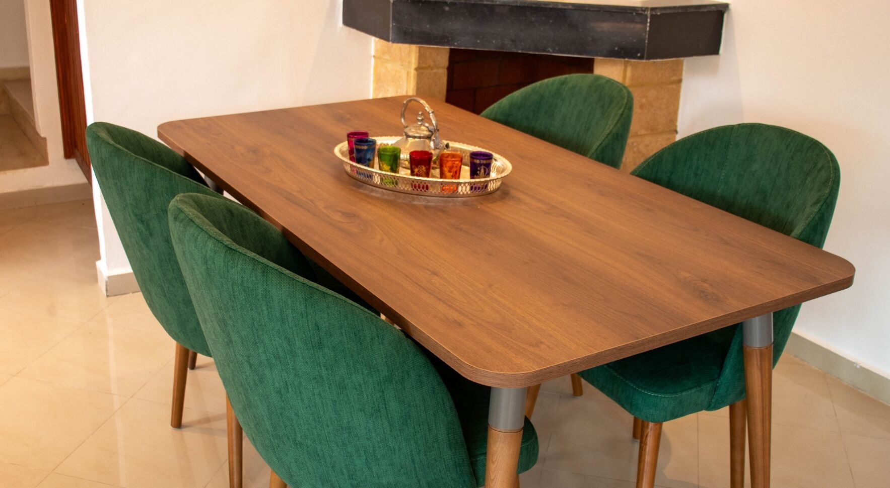 A dining table with a tray of colorful glasses and a teapot, surrounded by green upholstered chairs.