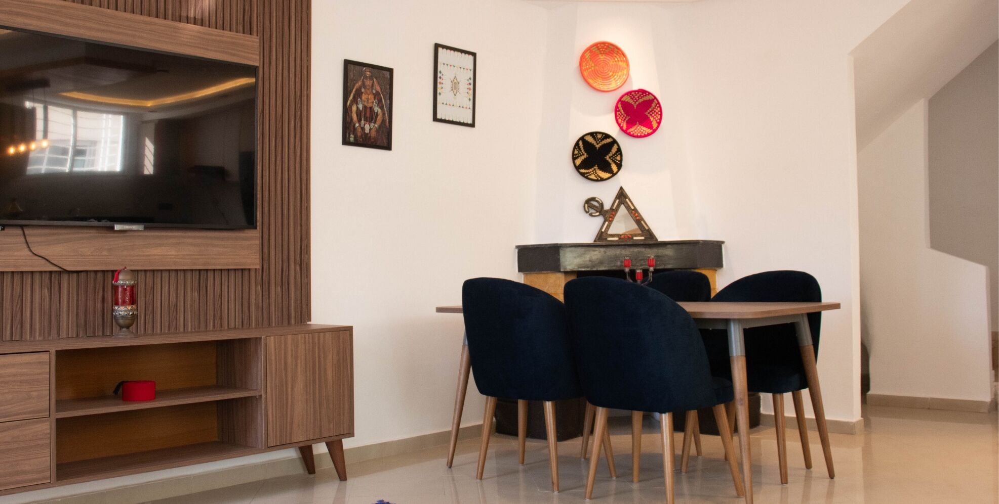 A modern living room with a wooden entertainment unit, a flat-screen TV, a dining table with four chairs, and decorative wall art.