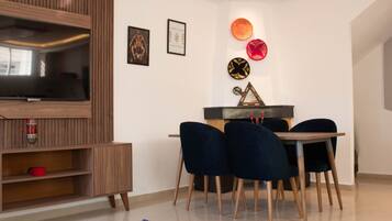A modern living room with a wooden entertainment unit, a flat-screen TV, a dining table with four chairs, and decorative wall art.