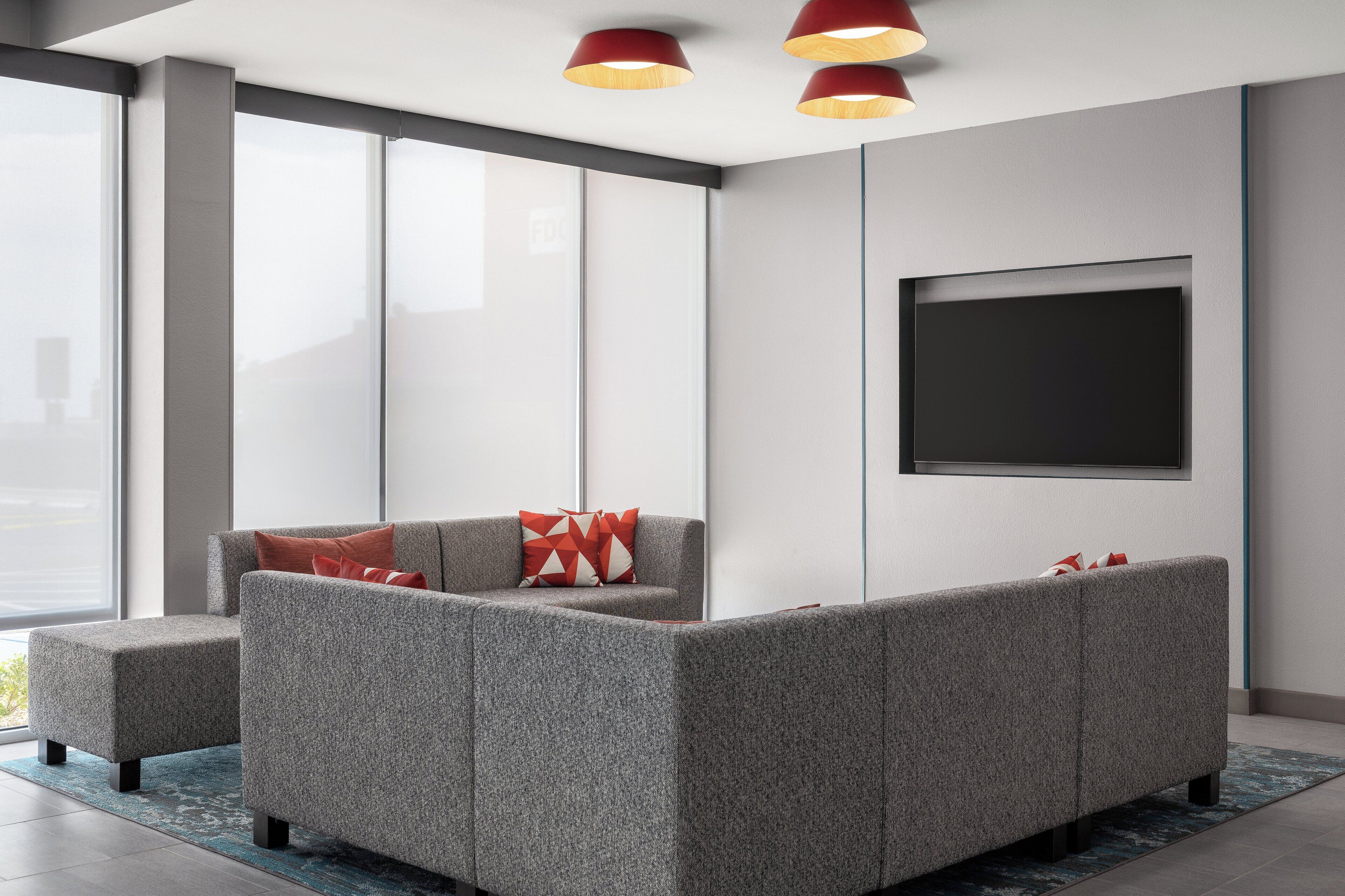 A modern living room with a grey sectional sofa, red cushions, and a large flat-screen TV mounted on the wall.