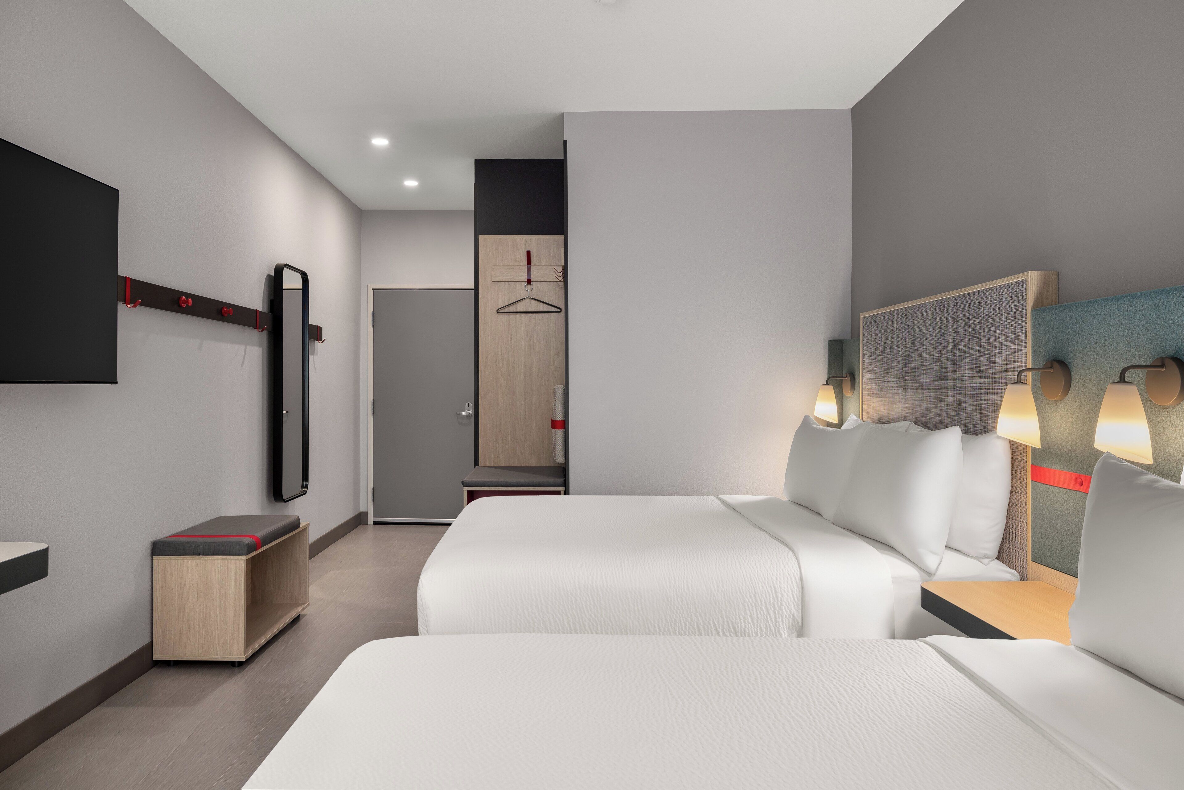 Una camera d'albergo moderna con due letti, una TV a schermo piatto, una panca in legno, un armadio e un bagno visibile attraverso una porta aperta.