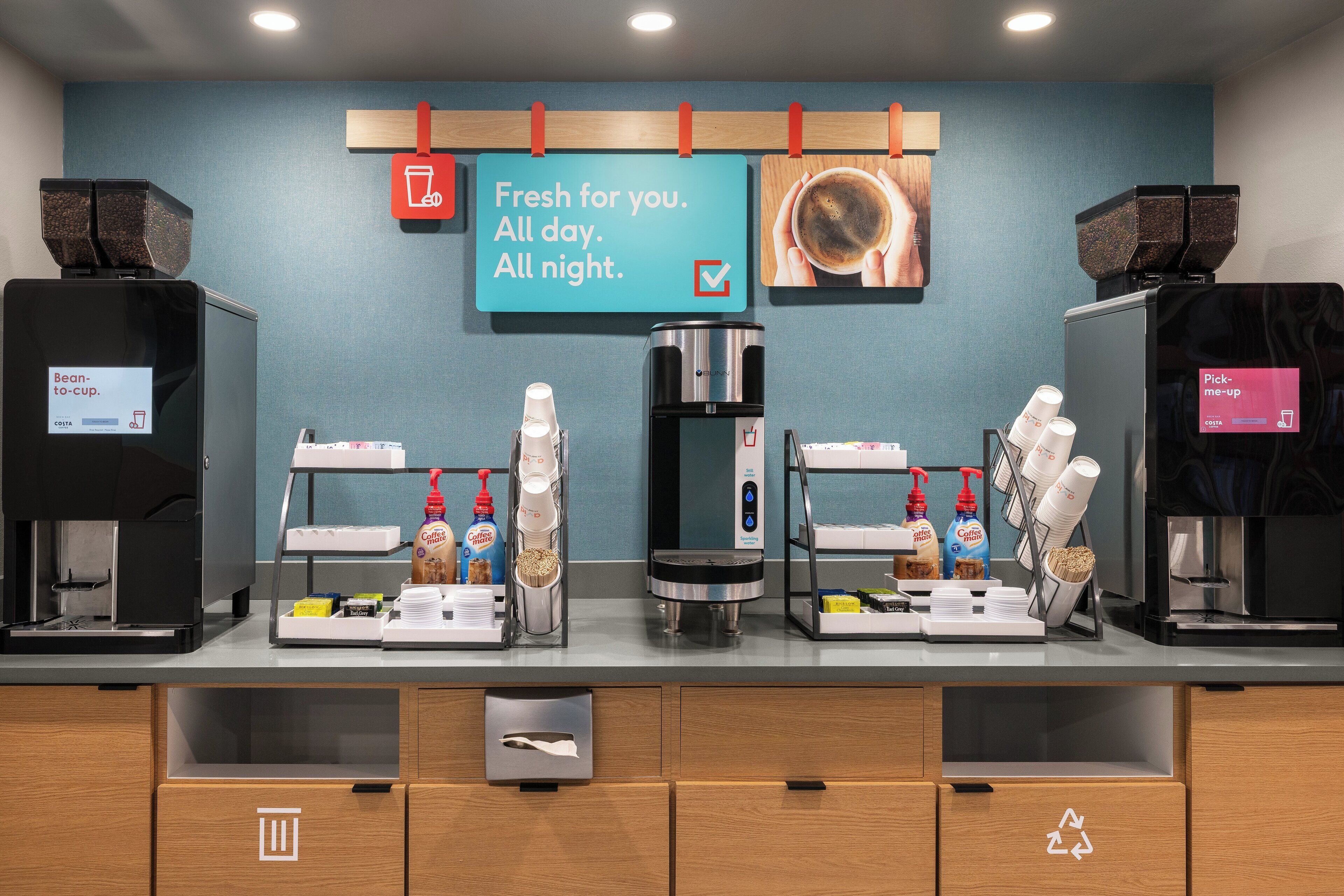 A coffee station with multiple coffee machines, a sign indicating availability of coffee all day and night, and a recycling bin.