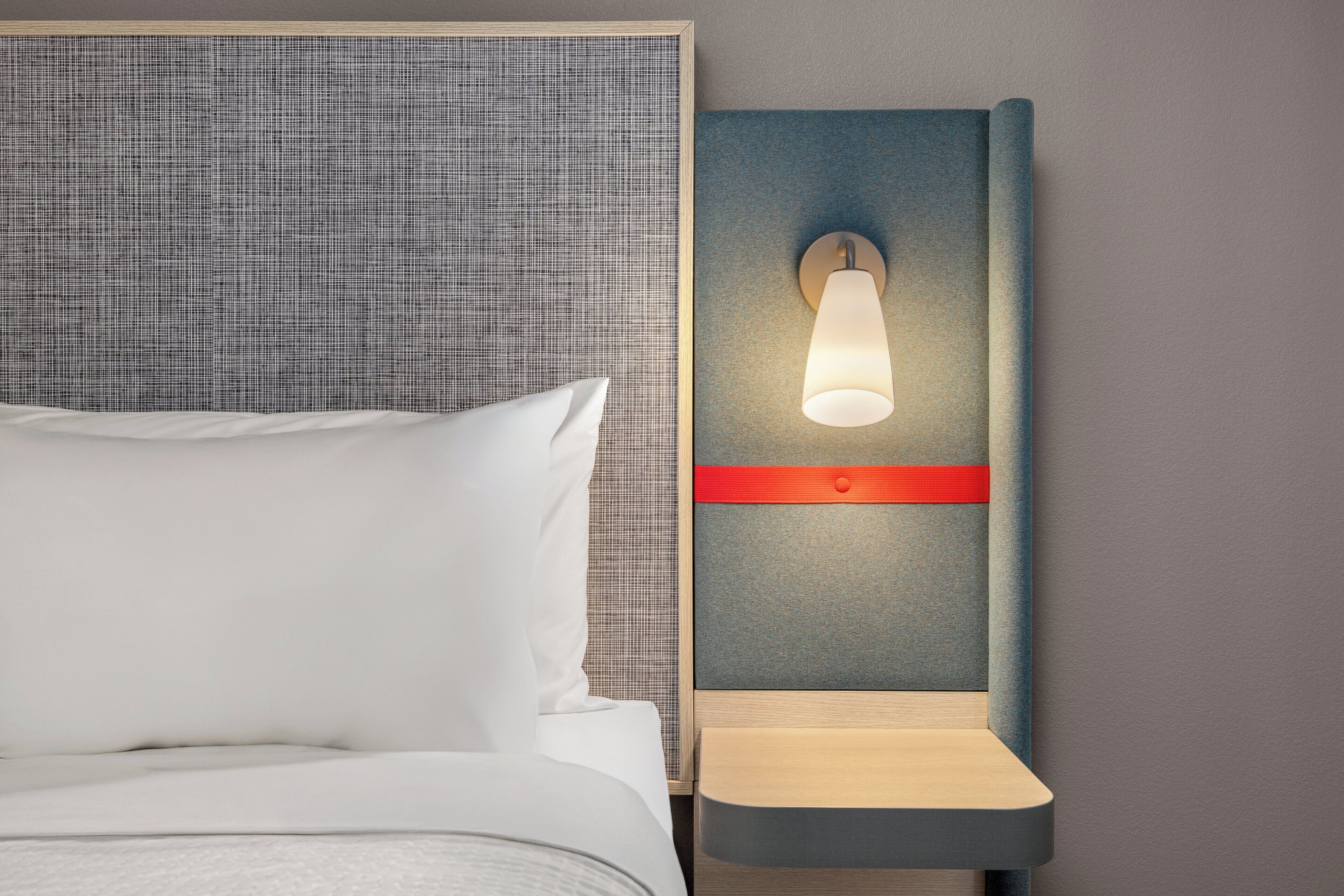 A neatly made bed with white linens and a pillow, a wall-mounted nightstand with a lamp, and a textured grey headboard.