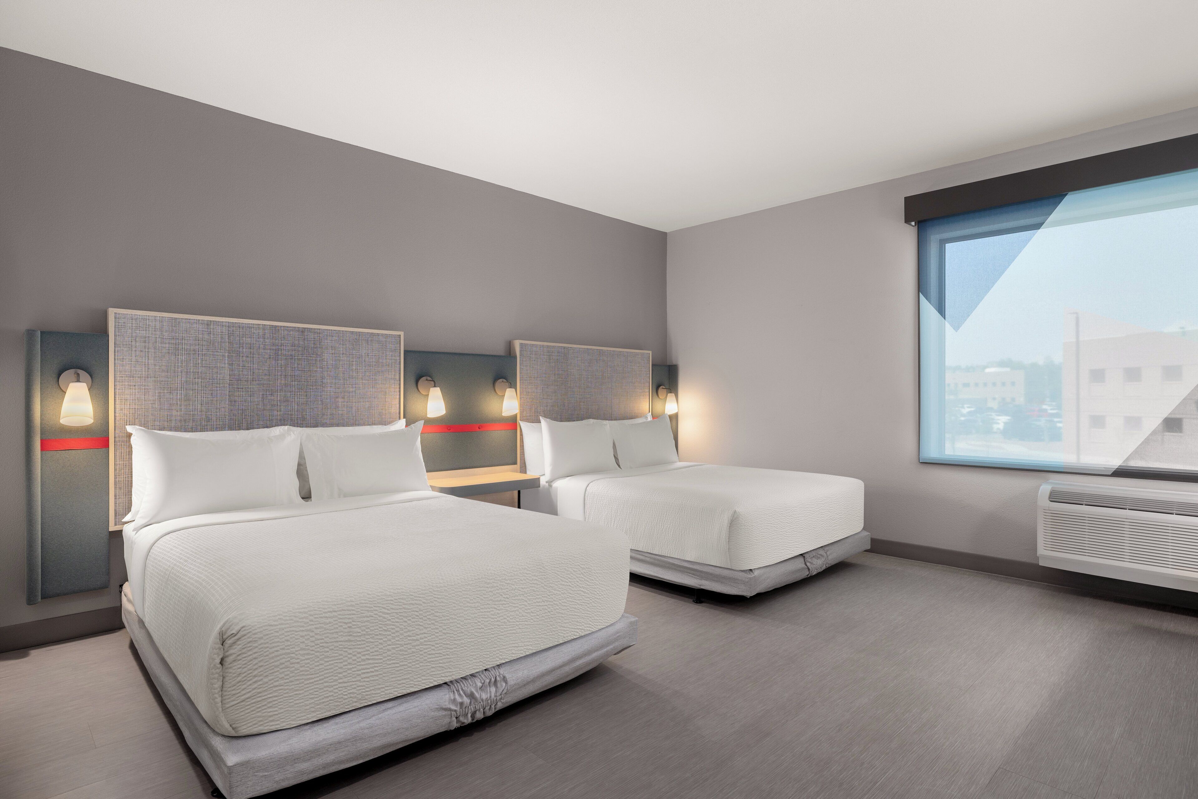 A modern hotel room with two beds, a large window, and a wall-mounted air conditioner.