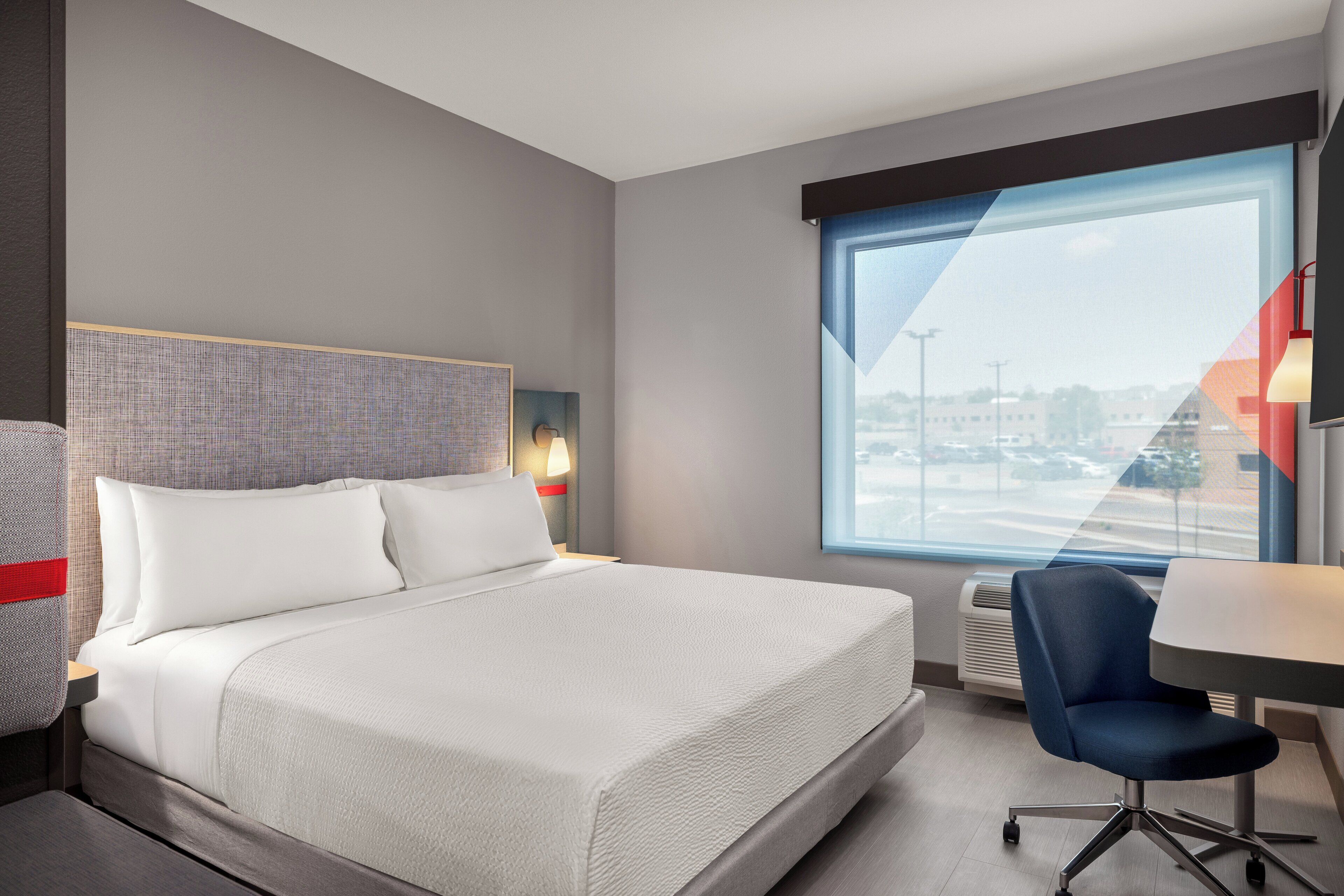 A hotel room with a large bed, a desk, and a chair. There is a window with a view of the city.