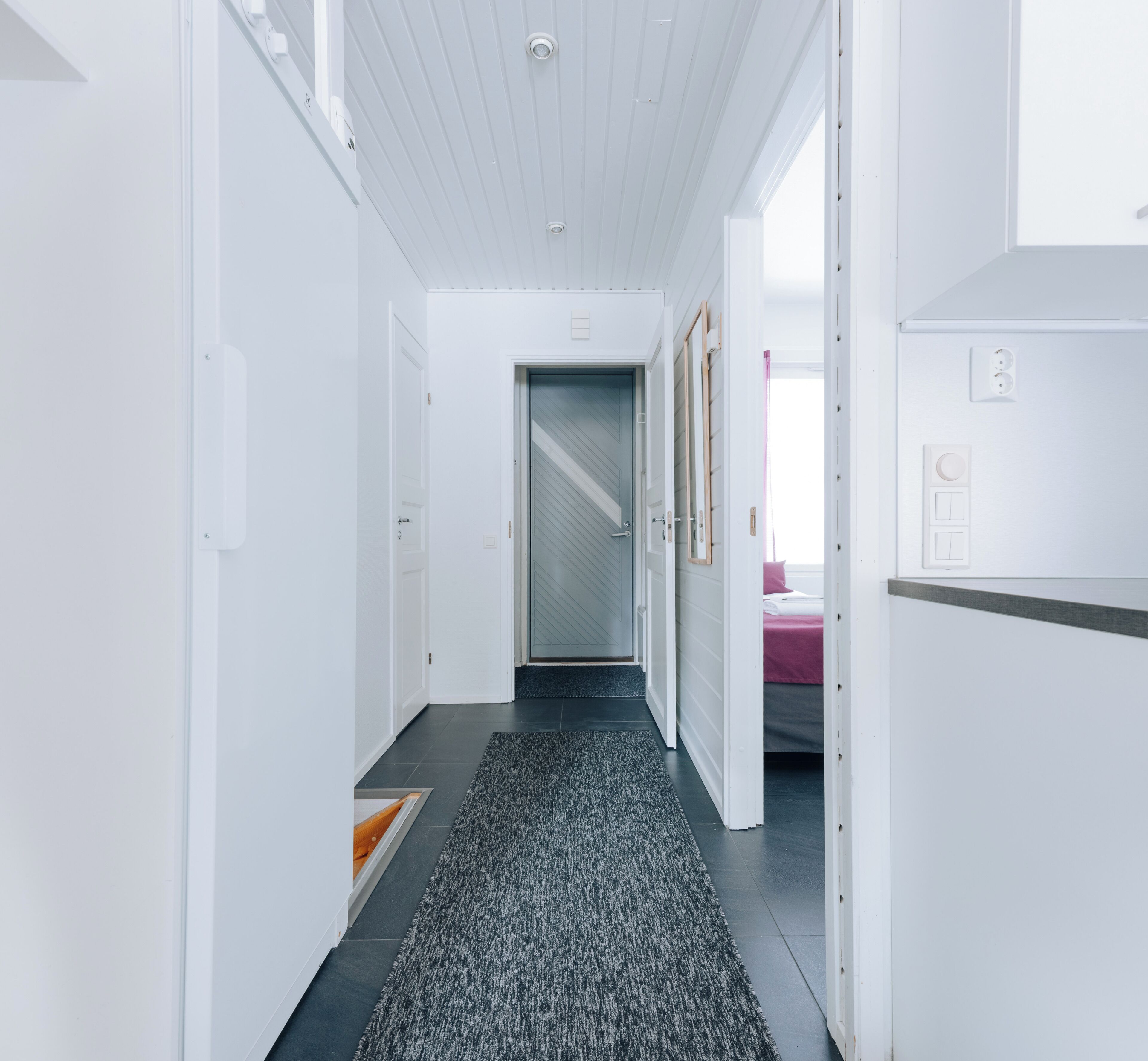 Un pasillo moderno con paredes blancas, alfombra gris y una puerta que da a una habitación con cama.