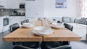 A modern kitchen with white cabinets, a wooden dining table set for six, and a sofa with patterned cushions in the background. - 3BR Holiday Apt. with Sauna (Kittilä)