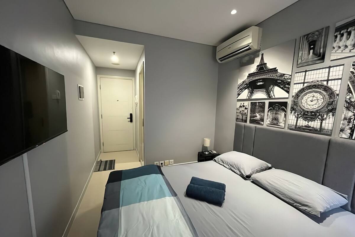 A modern bedroom with a bed, a flat-screen TV, a wall-mounted air conditioner, and a door leading to another room.