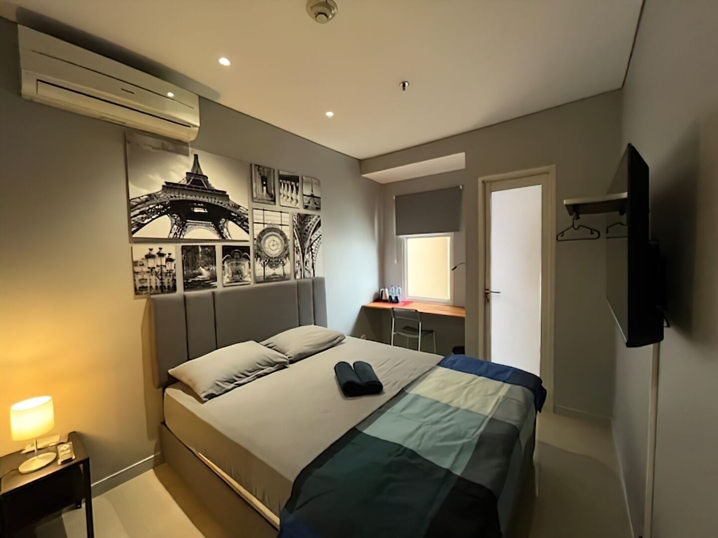 Une chambre à coucher moderne comprenant un lit, une table de chevet, un appareil de climatisation, un mur décoré d’œuvres d’art, un bureau avec une chaise, une fenêtre et un téléviseur fixé au mur.