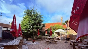 Eine Terrasse im Freien mit Holztischen und roten Sonnenschirmen, einem sternförmigen Tisch und einem Backsteinhaus im Hintergrund.
