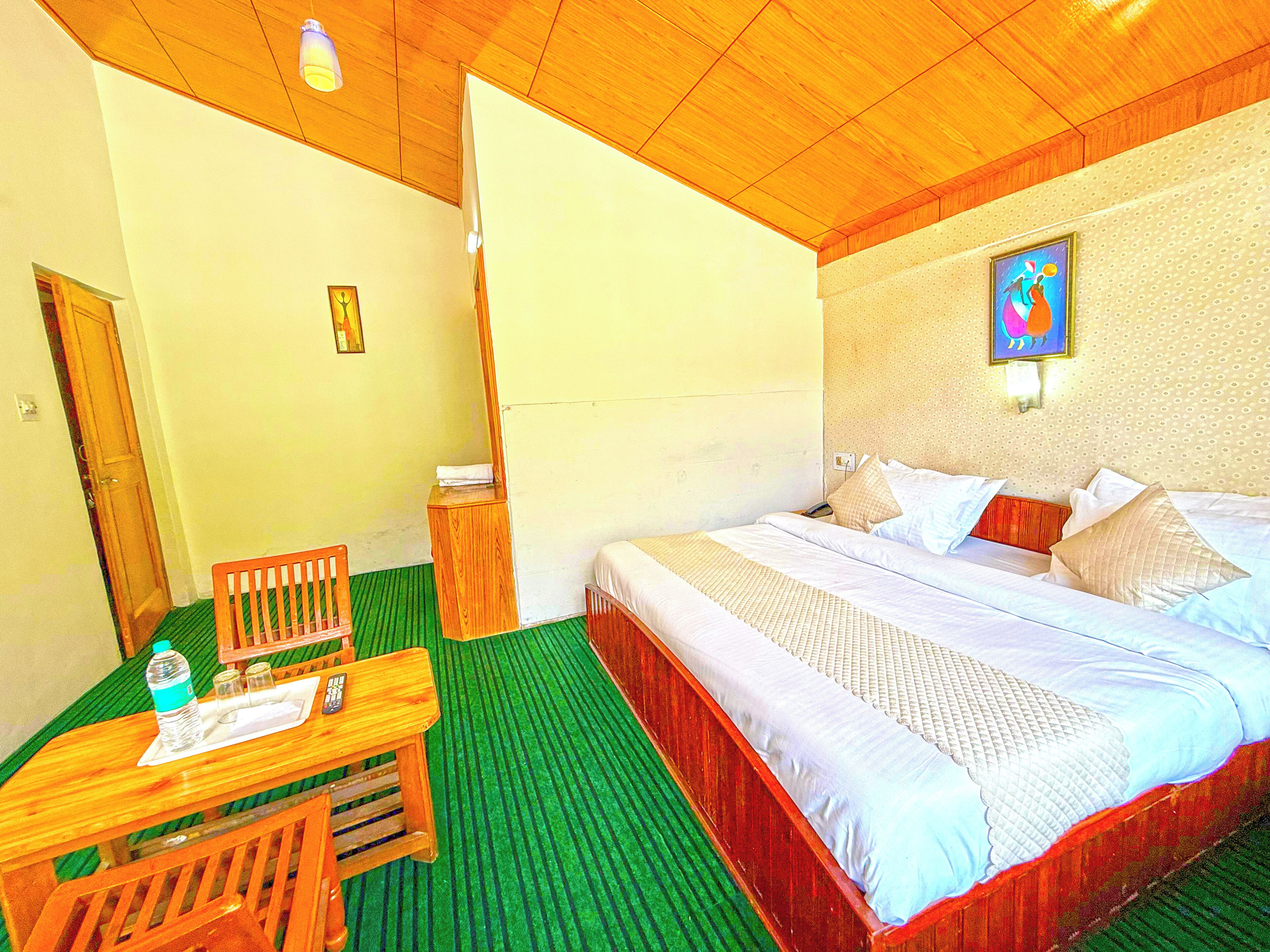 A hotel room with a bed, a desk with a chair, a wooden side table, a green carpet, and a wooden ceiling.
