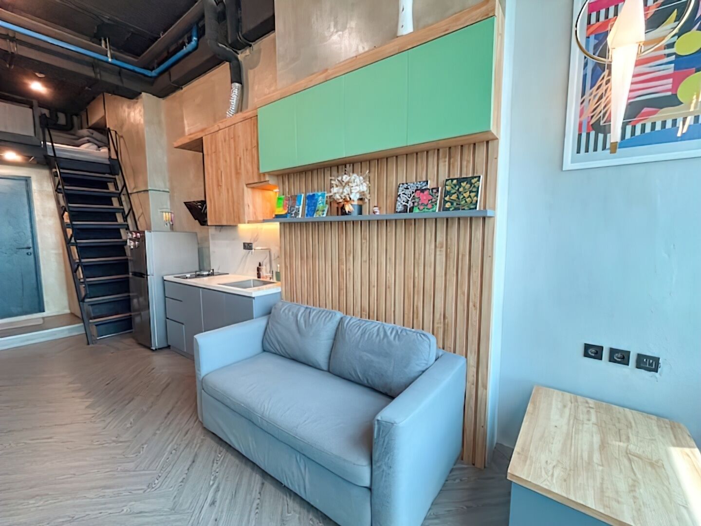 A modern living room with a grey sofa, wooden panel wall, and a kitchen area with a sink and cabinets.