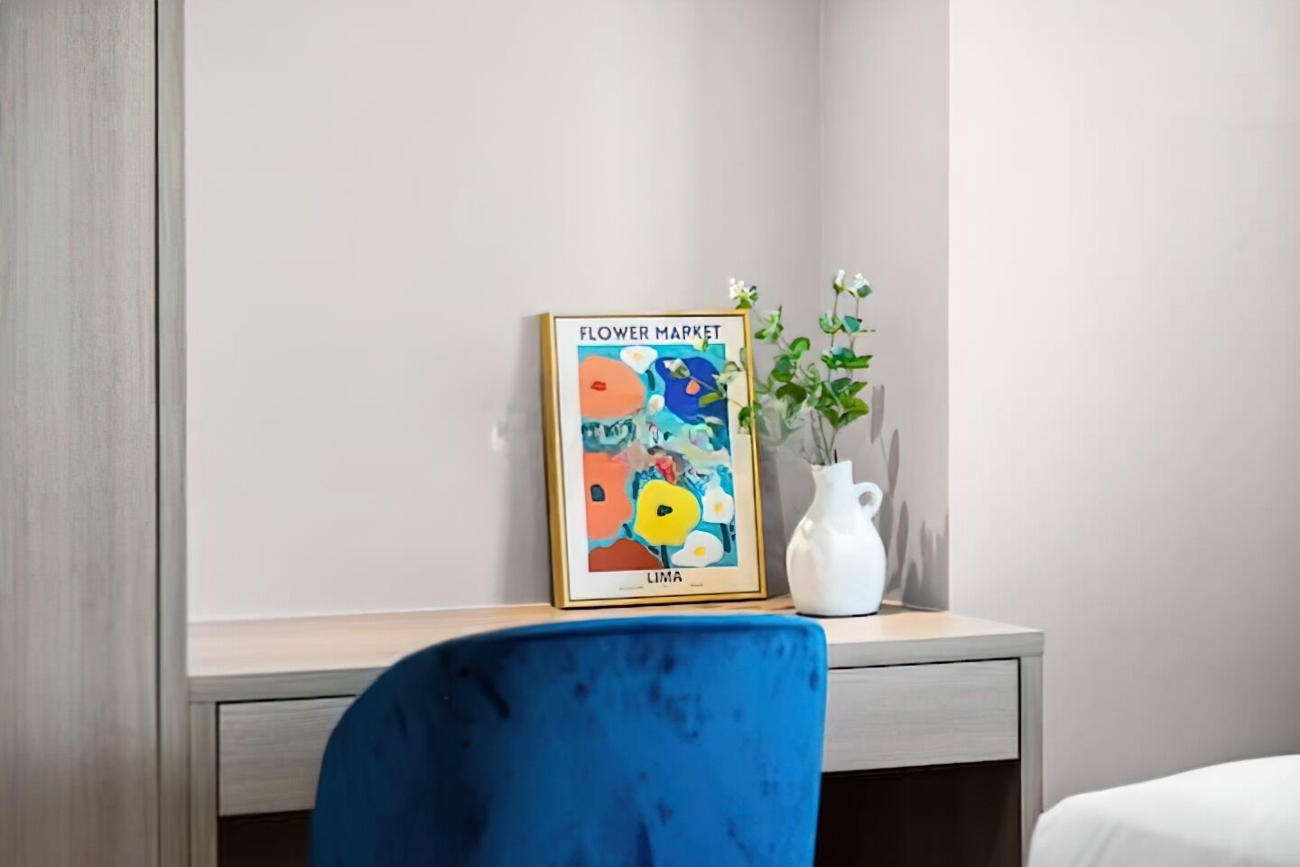 Un'opera incorniciata intitolata "Flower Market" di Lina su un comodino, un vaso bianco con piante verdi e una sedia blu.