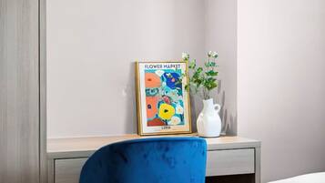Un'opera incorniciata intitolata "Flower Market" di Lina su un comodino, un vaso bianco con piante verdi e una sedia blu.