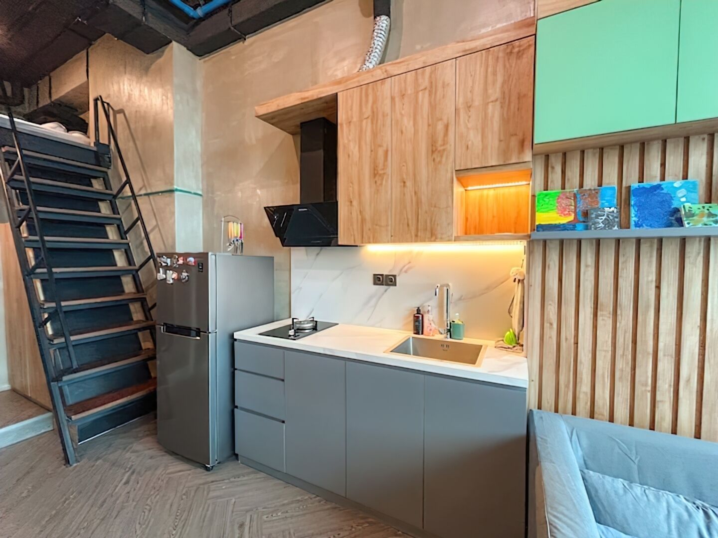 Cocina compacta con estufa integrada, lavamanos y refrigerador. Se observa un gabinete de madera con un estante sobre el lavamanos. Una escalera es visible a la izquierda.