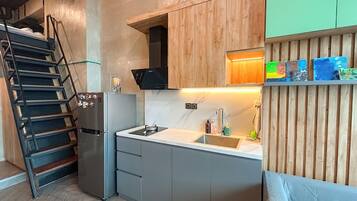 Cocina compacta con estufa integrada, lavamanos y refrigerador. Se observa un gabinete de madera con un estante sobre el lavamanos. Una escalera es visible a la izquierda.