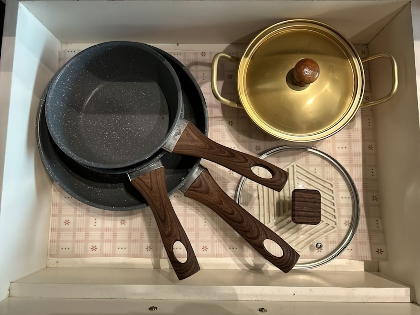 A kitchen drawer with two black frying pans, a glass lid, and wooden handles.