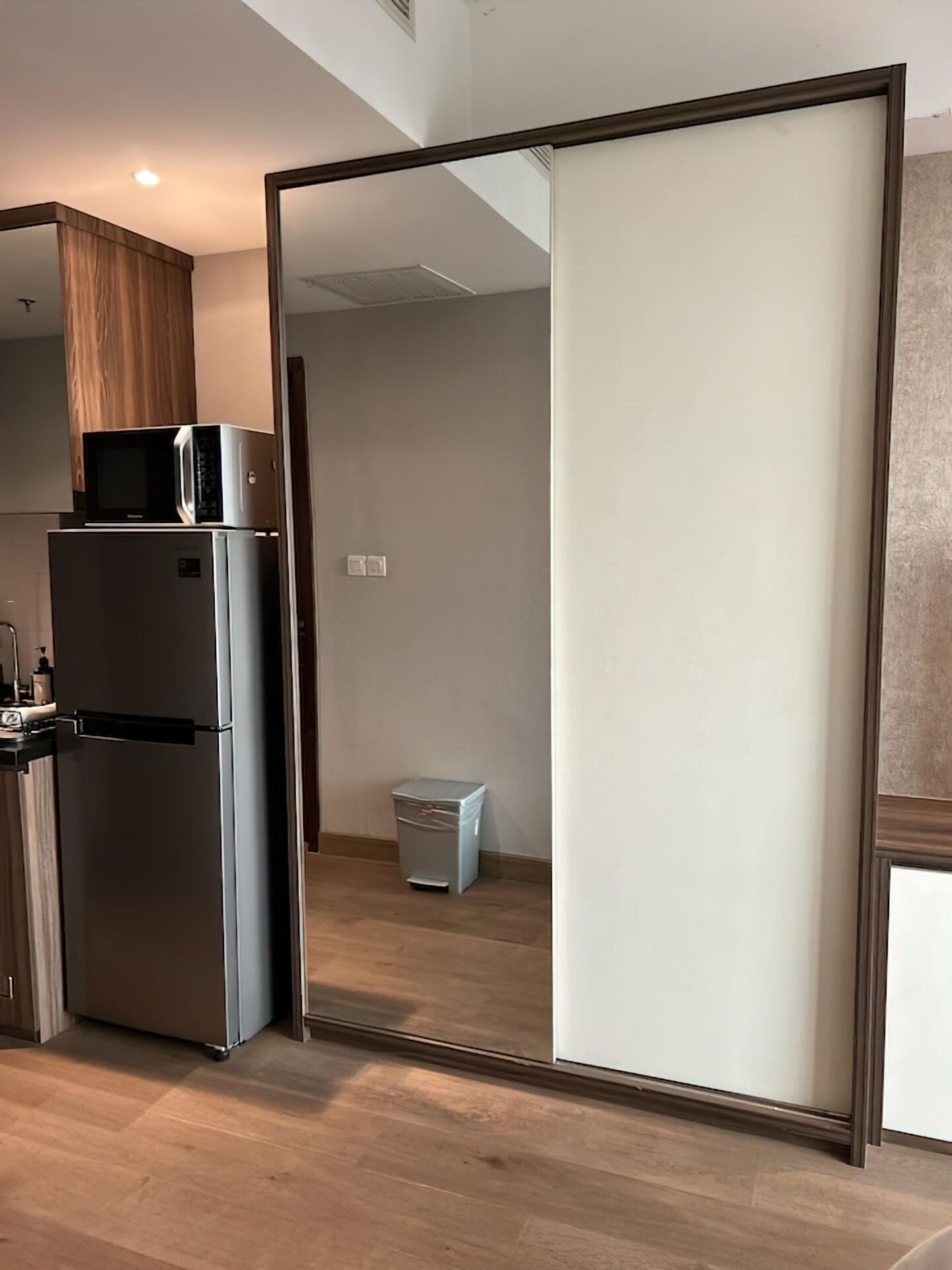 A modern kitchen with a built-in microwave and refrigerator, a sliding door leading to a bathroom, and a trash bin in the corner.