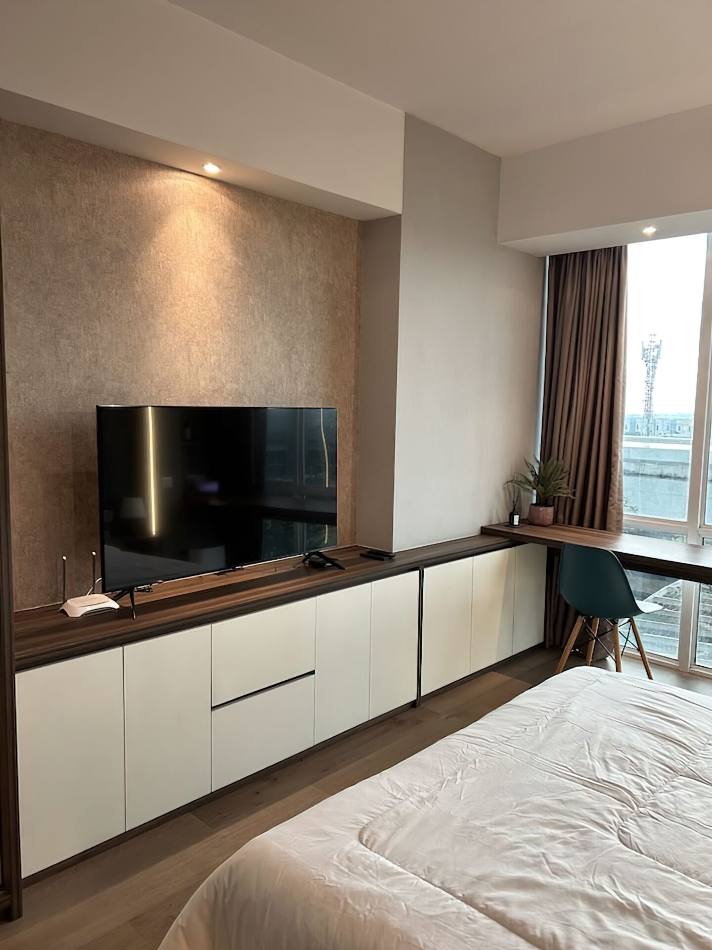 A modern hotel room with a large flat-screen TV mounted on a wooden wall, a desk with a chair, a bed with white bedding, and a view of a building through a window.