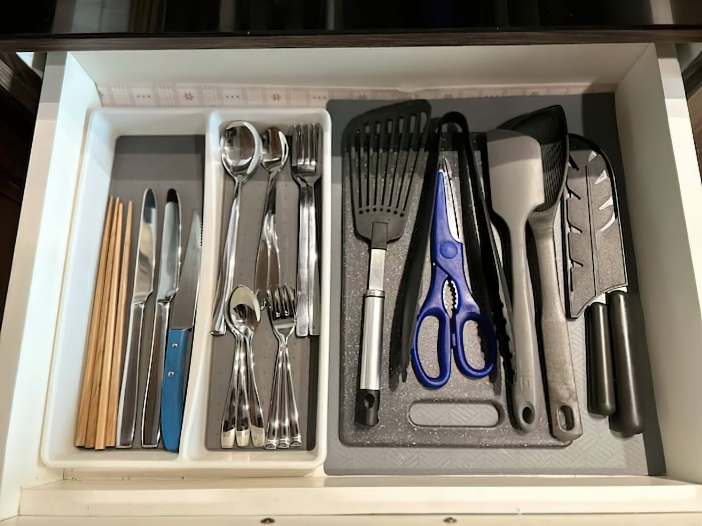 Un cassetto pieno di utensili e attrezzi da cucina, tra cui bacchette, coltelli, cucchiai, una spatola, pinze e forbici.