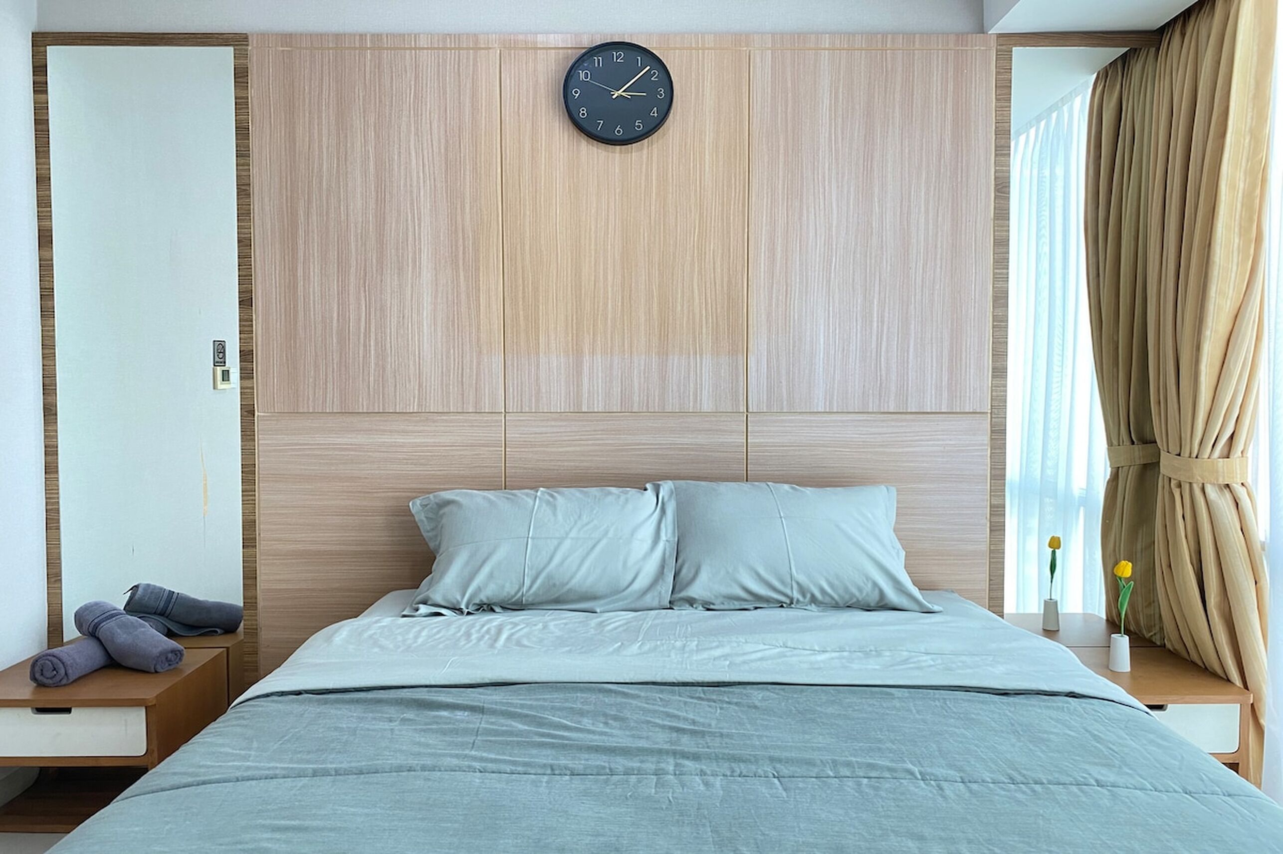 A modern bedroom with a wooden headboard, a clock, a bed with light-colored bedding, a nightstand with a lamp, and a window with curtains.