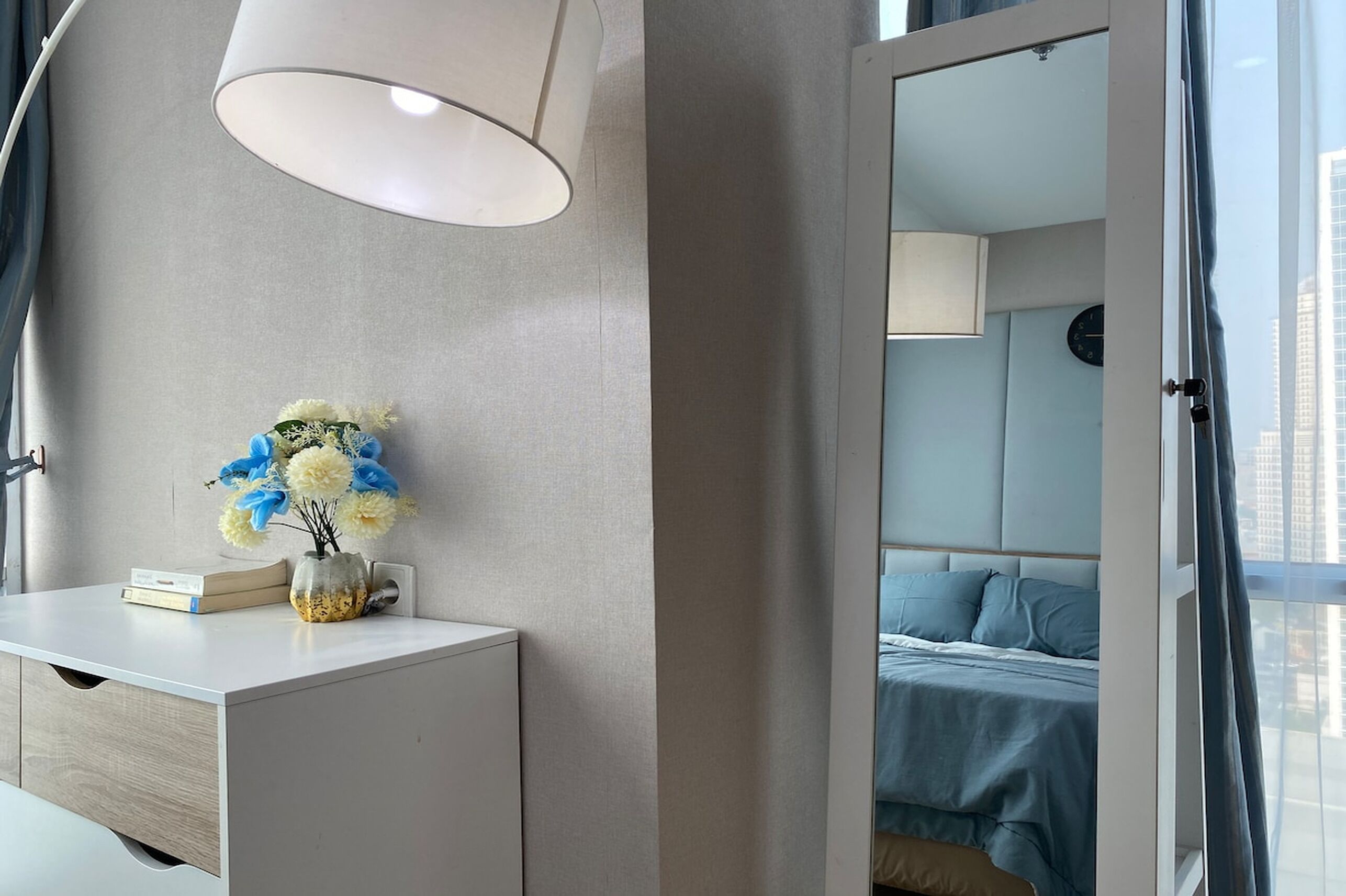 A modern hotel room with a white desk, a vase of flowers, a lamp, and a view of the city through a window.
