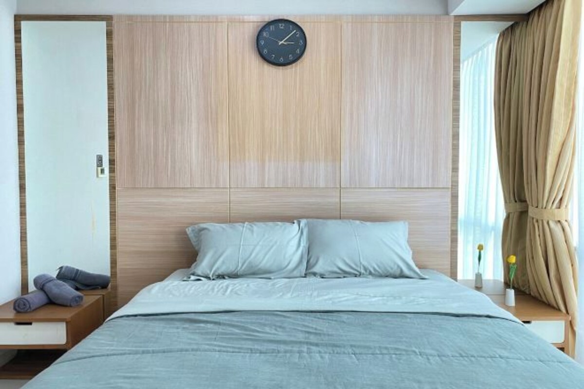 Un dormitorio moderno con cabecera de madera, un reloj, una cama con ropa de cama en tonos claros y una mesita de noche con una lámpara y un jarrón de flores.