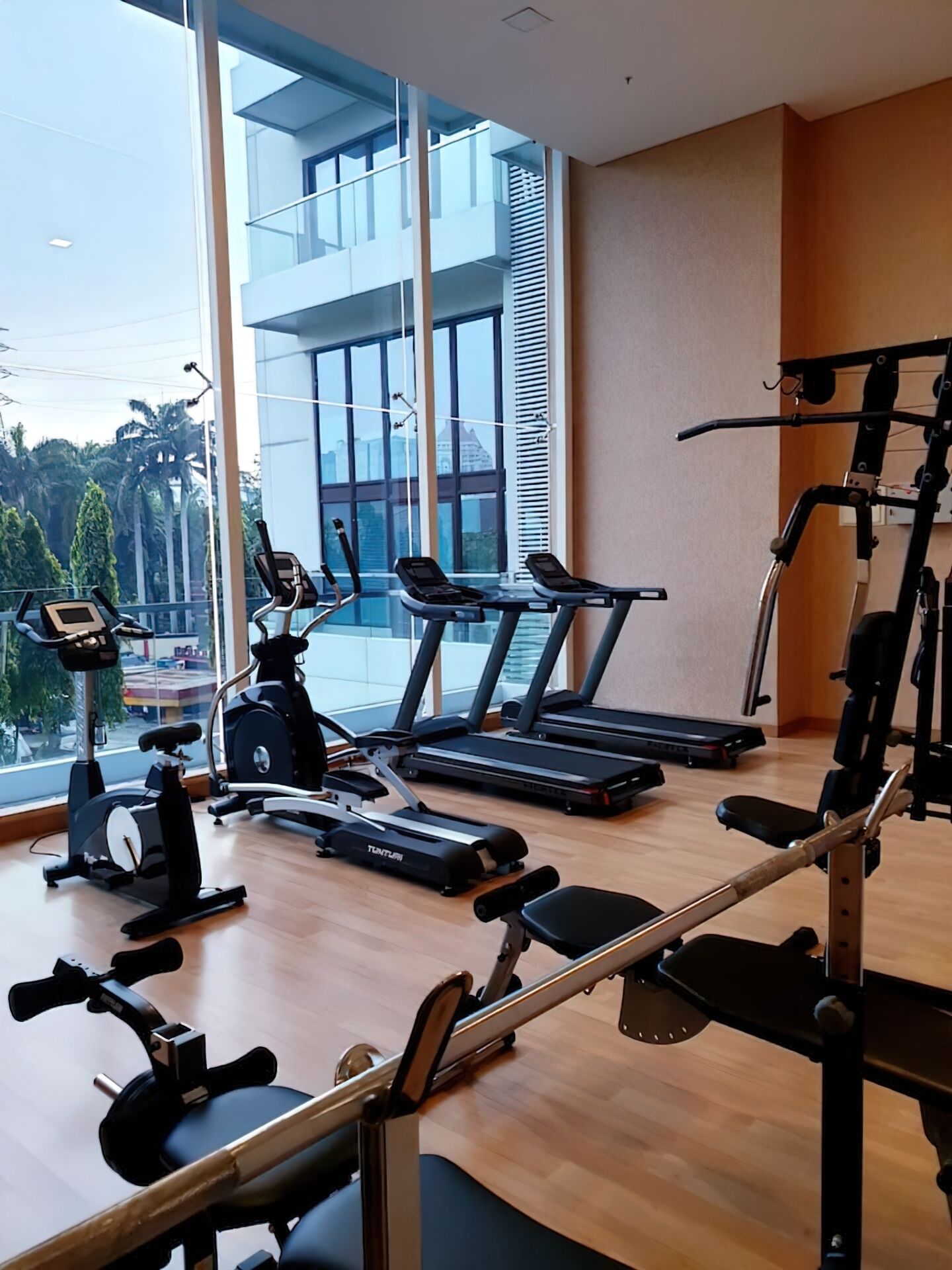 Une salle de sport moderne équipée de tapis de course, de vélos d’appartement et d’appareils de musculation. De grandes fenêtres offrent une vue sur un immeuble et des palmiers.