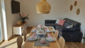 A dining area with a wooden table set for four, a dark grey sofa, and a wall-mounted TV.