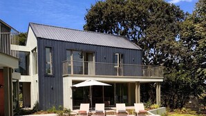 A modern two-story house with a swimming pool, outdoor seating, and a large tree in the background. - New, stylish, 3 bedroom home, with infinity pool and Table Mountain views (Cape Town)