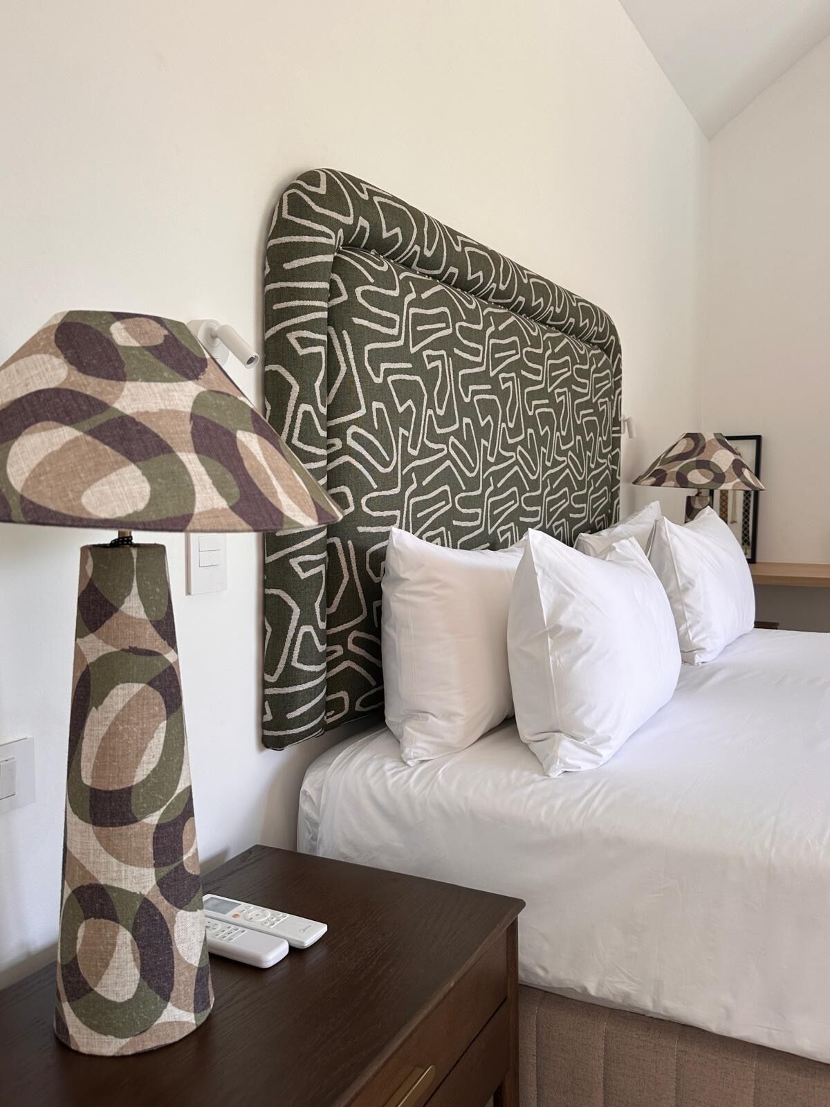 A hotel room with a patterned headboard, white bedding, a bedside lamp, and a remote control on the nightstand.