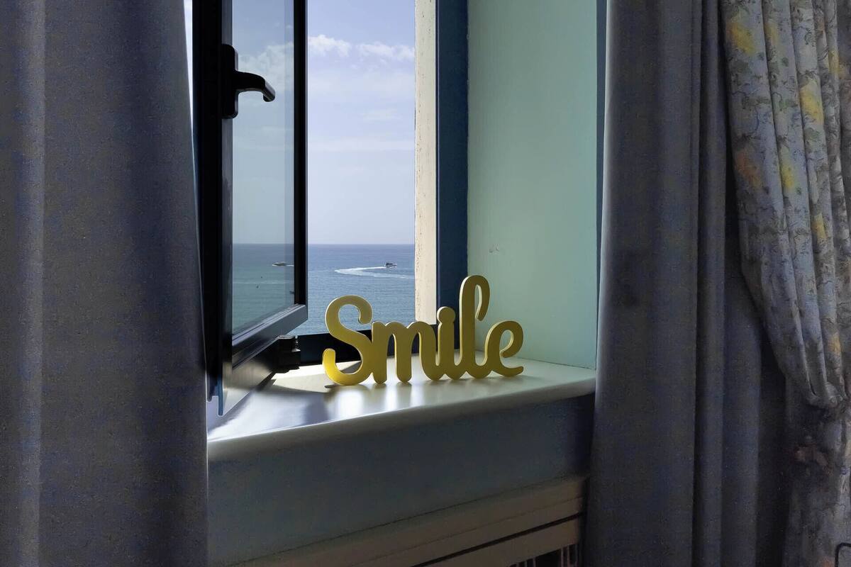 Una finestra con vista sull'oceano, tende e una decorazione con la scritta 'Smile' sul davanzale.