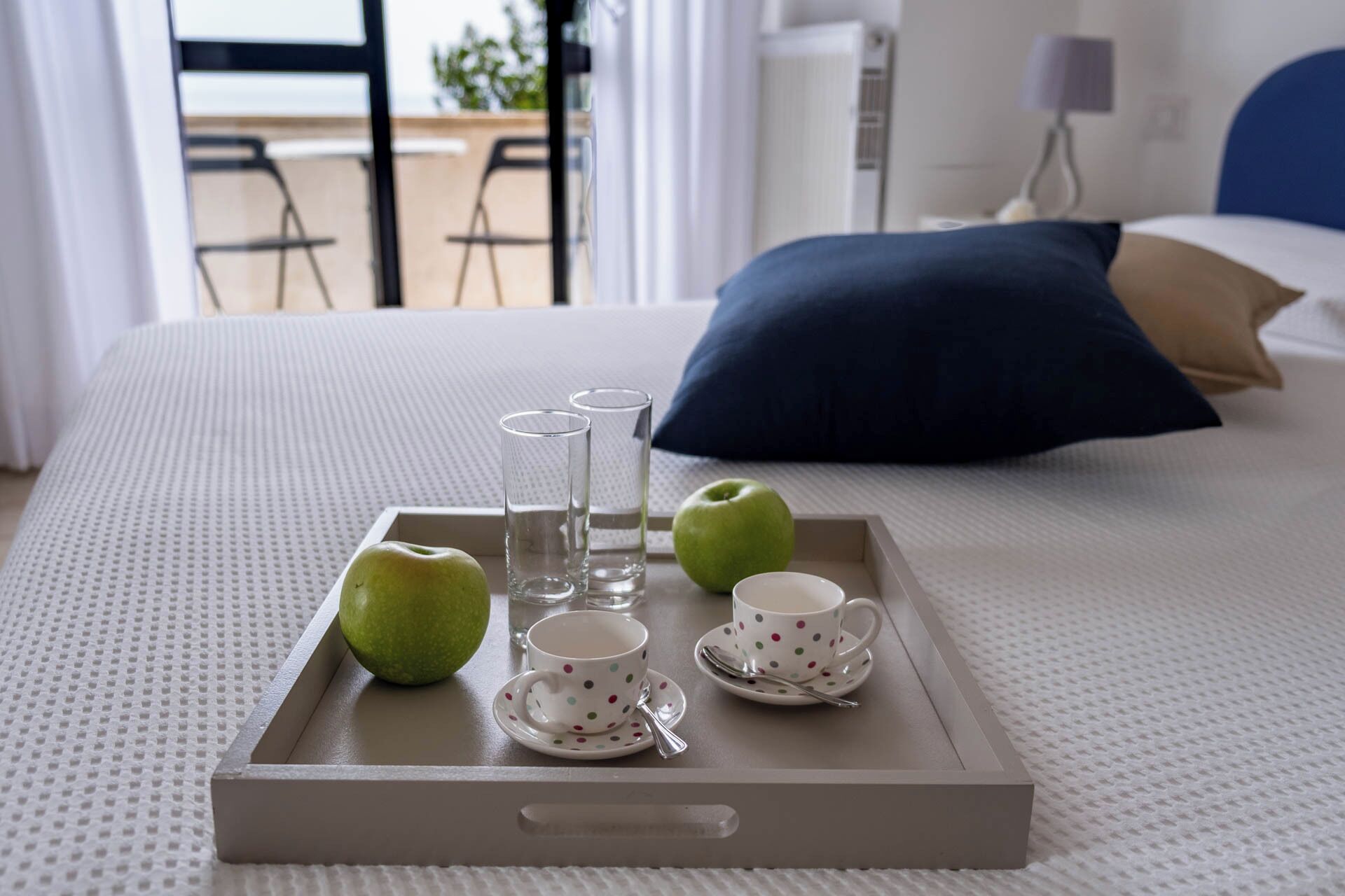 A neatly arranged breakfast tray with apples, glasses, and a cup on a bed.
