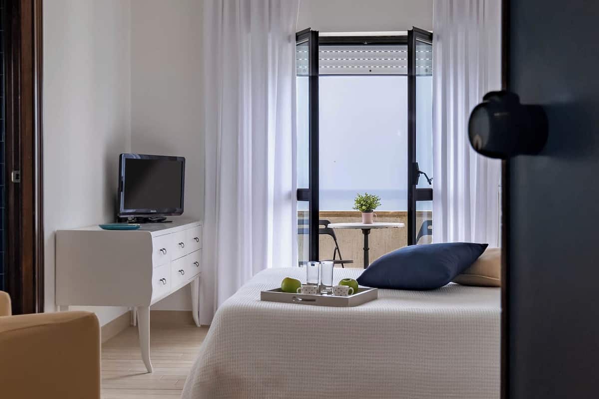 Una camera d'hotel con un letto, una scrivania con la televisione, un balcone con vista e una finestra con tende.