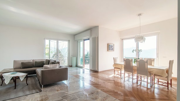 A spacious living room with a sofa, a dining table with chairs, and a view of the outdoors through large windows.