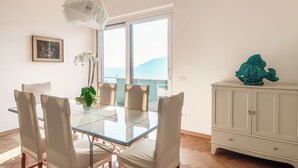 A dining area with a glass table, white chairs, a white cabinet, and a view of the sea.