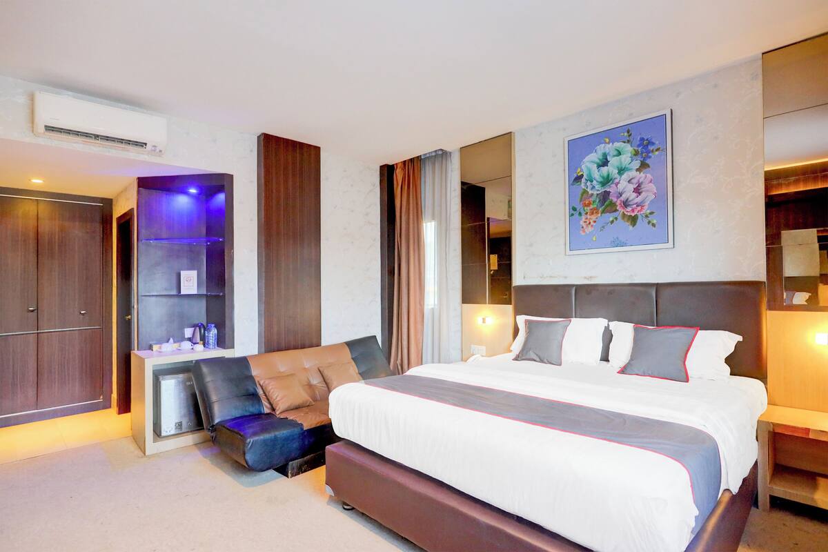 A modern hotel room with a large bed, a seating area with a sofa and ottoman, a built-in wardrobe, and a wall-mounted air conditioning unit.